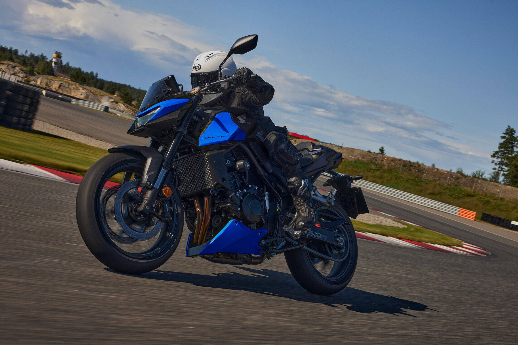 Honda CB750 Hornet 2027 in Glint Wave Blue in Schräglage auf einer Rennstrecke, Fahrer mit weißem Helm und schwarzer Lederkombi, Frontansicht von schräg links