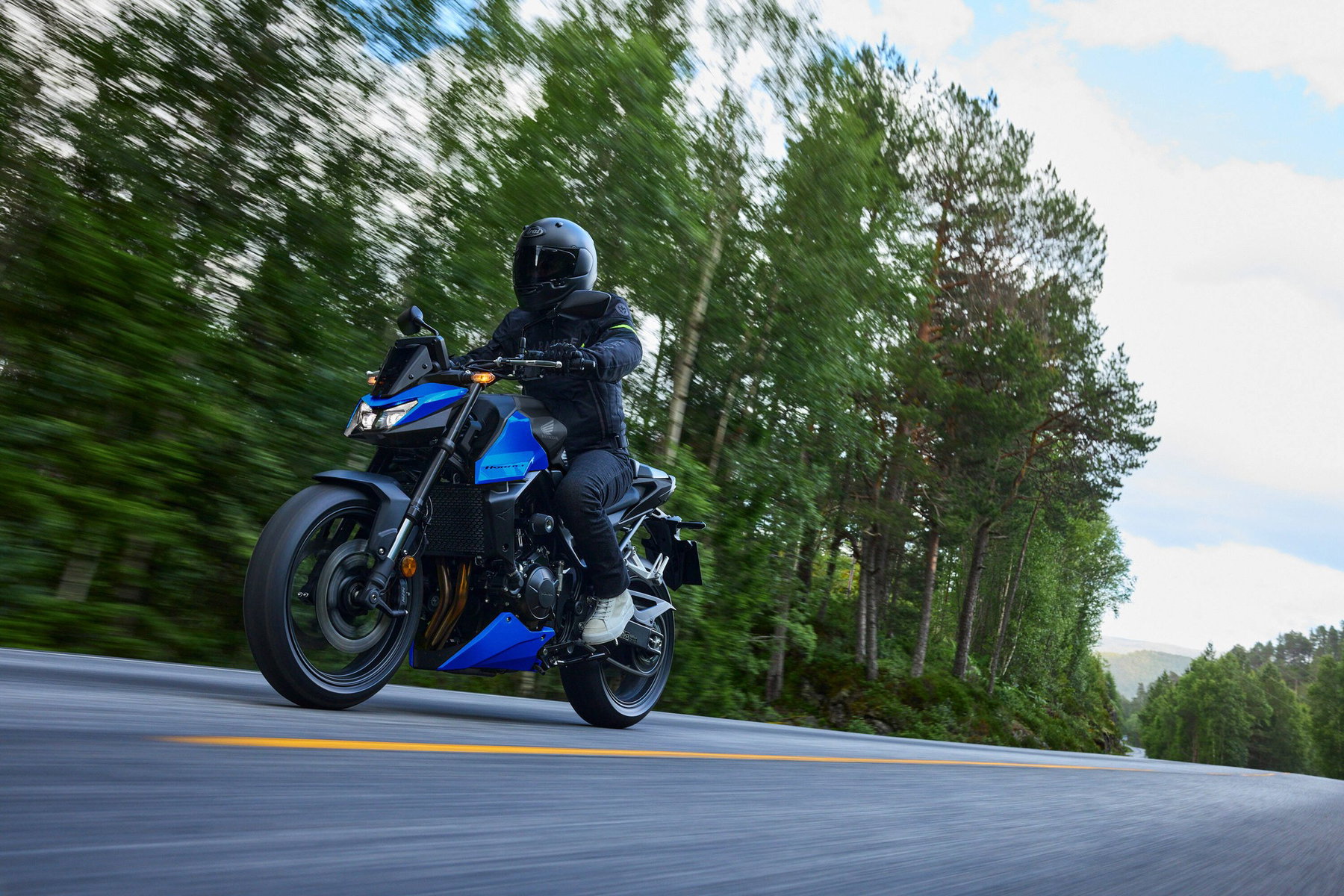 Honda CB750 Hornet 2027 in Glint Wave Blue auf einer Landstraße im Wald, Fahrer in Alltagskleidung, Frontansicht von schräg links in Bewegung