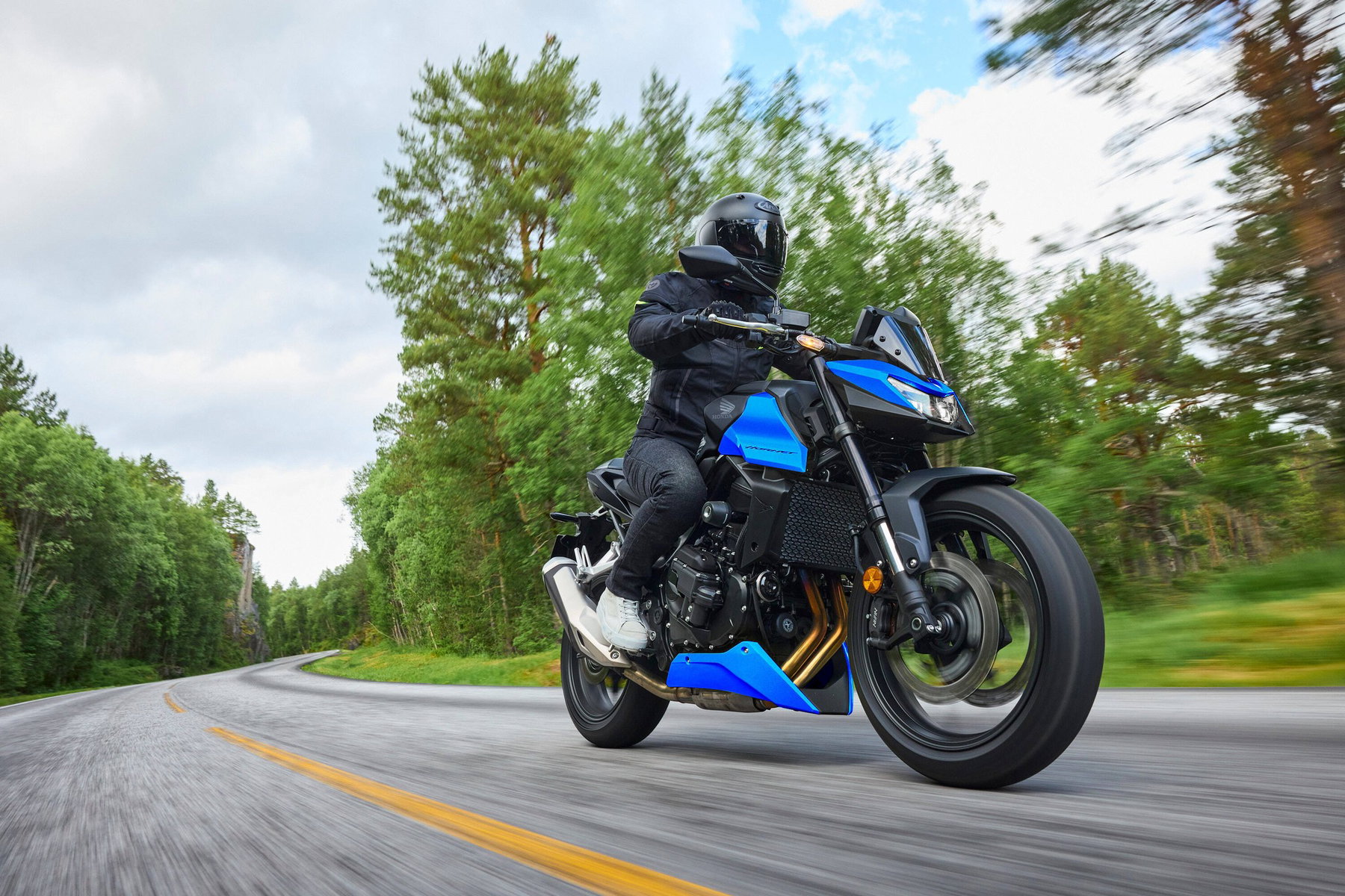 Honda CB750 Hornet 2027 in Glint Wave Blue in einer Landstraßenkurve, Frontansicht mit sichtbarem Kühler, Bugspoiler und Upside-down-Gabel