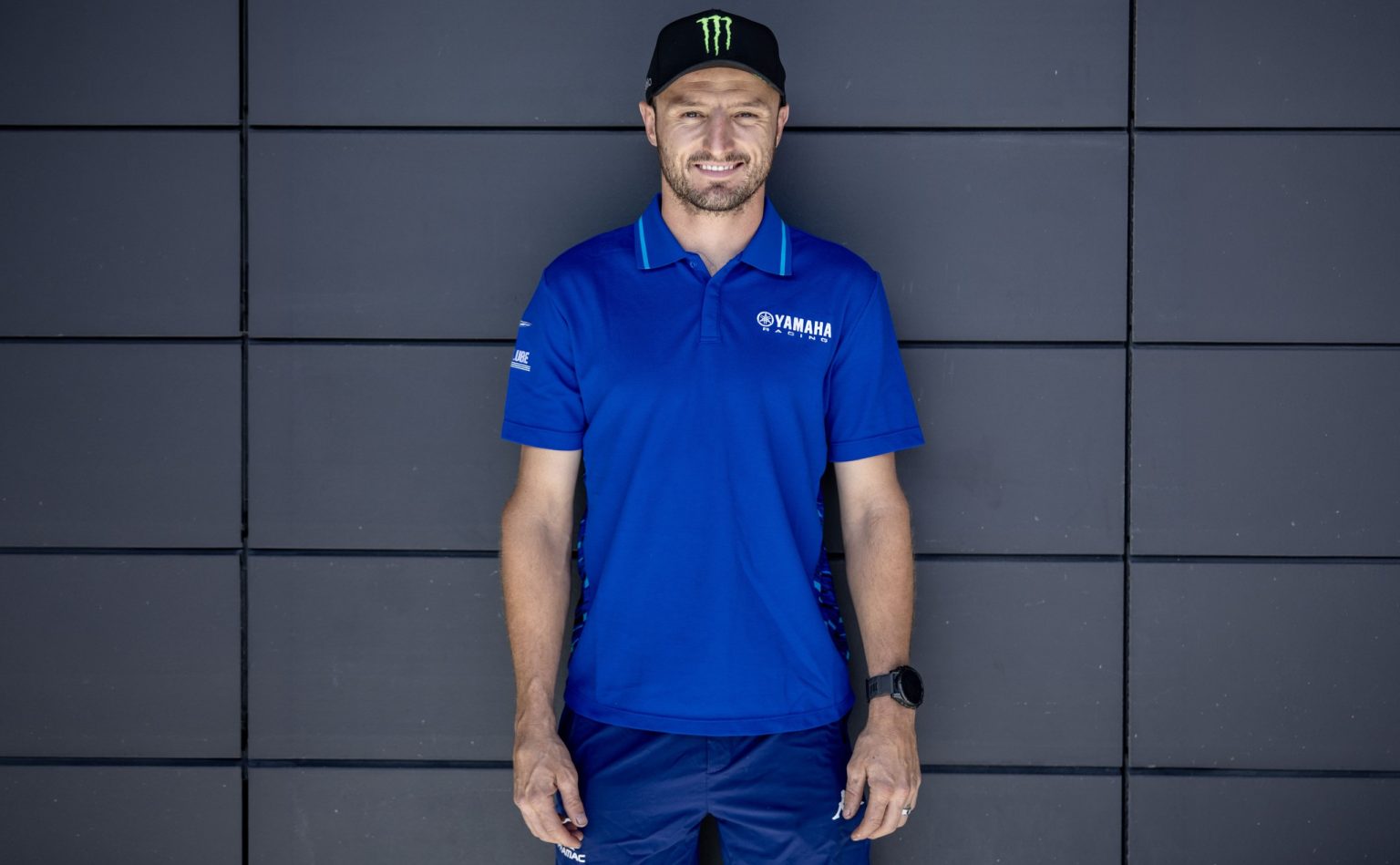 Jack Miller Leaves MotoGP: Yamaha Signs Him for World Superbike Jack Miller im blauen Yamaha-Racing-Polohemd und schwarzer Monster-Energy-Cap vor einer dunkelgrauen Paneelwand – Querformat-Ankündigungsbild zum Wechsel in die Superbike-WM 2027