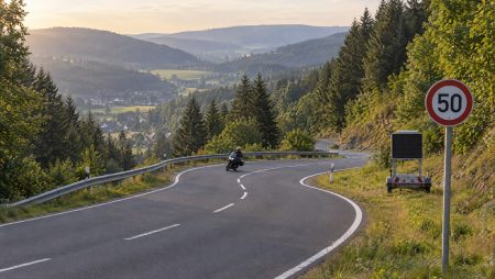 Motorradfahrer in einer Kurve einer bewaldeten Serpentinenstraße im Sauerland, am Straßenrand ein Tempo-50-Schild und eine mobile Geschwindigkeitsanzeigetafel (KI-generiertes Symbolbild)