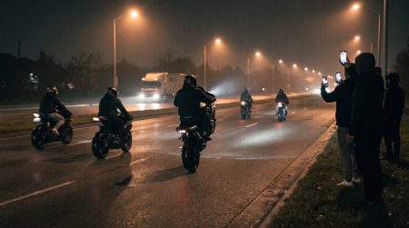 Illegale Motorradrennen Piacenza: mehrere Sportmotorräder rasen nachts über eine neblige Ausfallstraße am Stadtrand, Zuschauer filmen mit Smartphones am Straßenrand, im Hintergrund nähert sich ein Lkw.