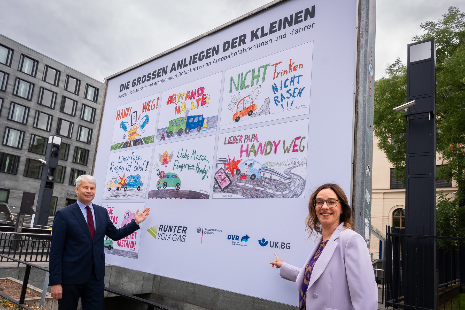 Runter vom Gas: Kinderbilder statt Leichensäcke an der Autobahn 1 Steffen Bilger und Christiane Leonard zeigen auf die große Plakatwand „Die großen Anliegen der Kleinen“ mit sieben Kinderzeichnungen der Kampagne „Runter vom Gas“