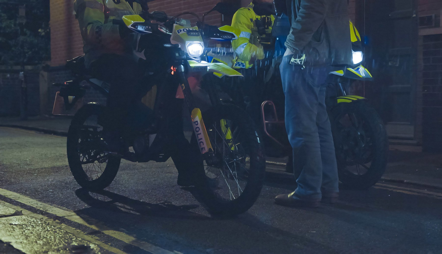 London Police Ride the Sur-Ron Ultra Bee: 25 Bikes Against Phone Snatching 3 Nahaufnahme mehrerer geparkter, gelb-schwarz lackierter Sur-Ron-Ultra-Bee-Polizeimotorraeder mit Aufschrift Police bei Regen und Dunkelheit, im Hintergrund Beamte im Einsatz