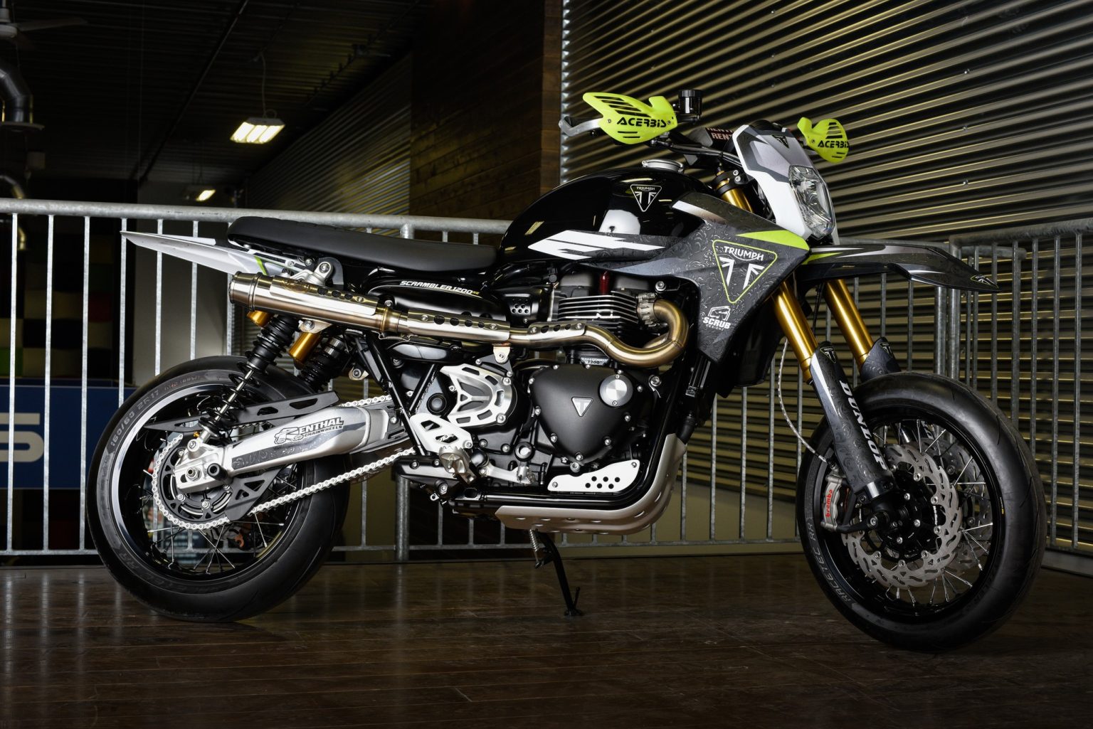 Triumph Originals 2026: Wie sieben Werkstätten die Scrambler neu denken Triumph Scrambler 1200 Custom-Umbau aus Kanada – Supermoto-Umbau in Schwarz-Grau mit gelben Acerbis-Handprotektoren, goldener Gabel und hochgelegter Auspuffanlage in einer Halle