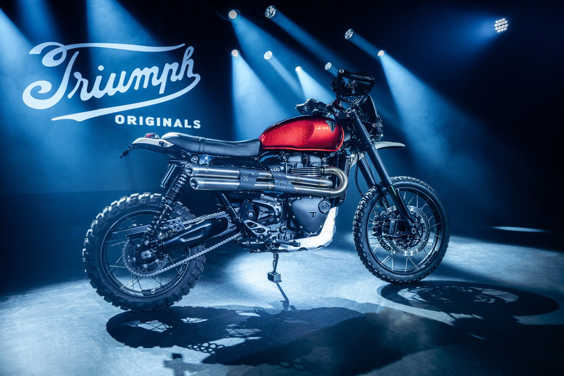 Triumph Originals 2026: Wie sieben Werkstätten die Scrambler neu denken 1 Triumph Scrambler 1200 Custom-Umbau aus Frankreich – Scrambler mit rotem Metallflake-Tank und Doppelauspuff im Scheinwerferlicht vor Triumph-Originals-Logo