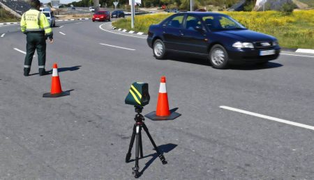 Guardia-Civil-Beamter in gelber Warnweste steht an einer Straßenkreuzung neben einem auf Stativ montierten, kompakten schwarz-gelben Veloláser-Radargerät, im Hintergrund ein dunkler Pkw und Warnkegel auf der Fahrbahn