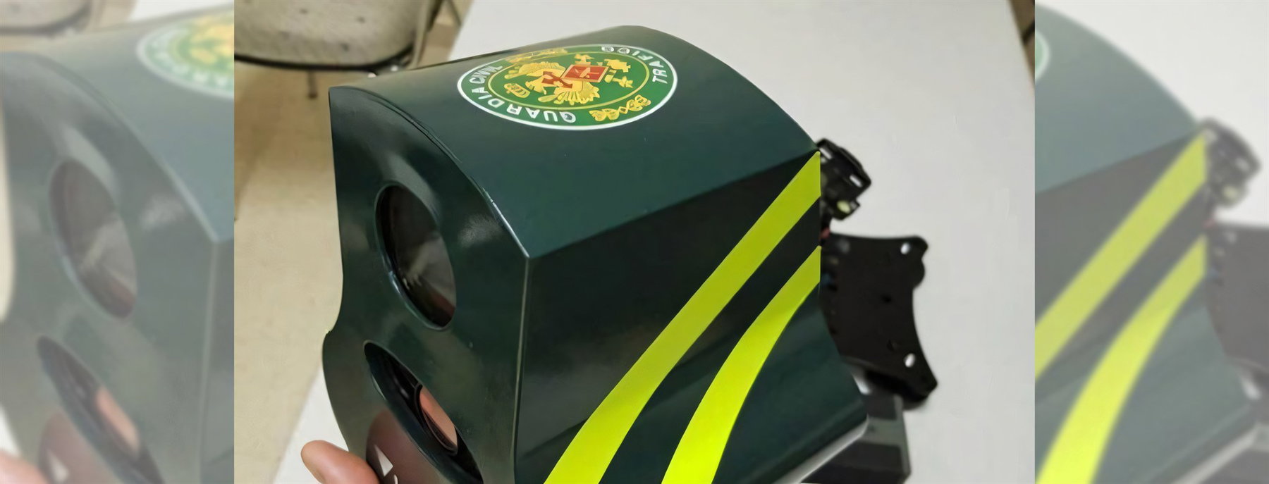 Detailaufnahme eines dunkelgrünen Veloláser-Gehäuses mit gelben Streifen und rundem Guardia-Civil-Wappen, in der Hand gehalten auf einem Tisch, im Hintergrund unscharf ein Stuhl