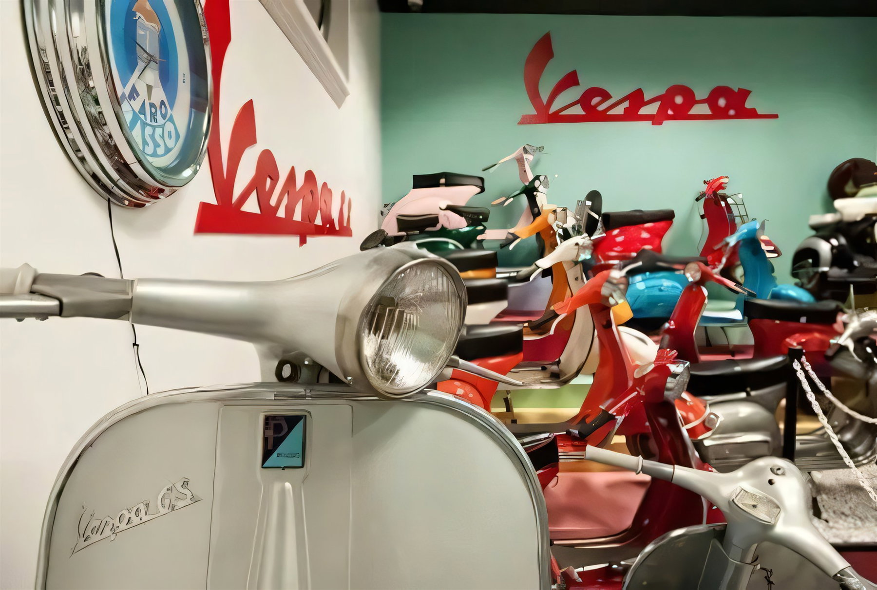 Vespa Primavera wird zur EU-Marke: Was das für Retro-Roller bedeutet 4 Klassische Vespa-Roller mit Chromscheinwerfer vor roten Vespa-Schriftzügen in einer Ausstellung