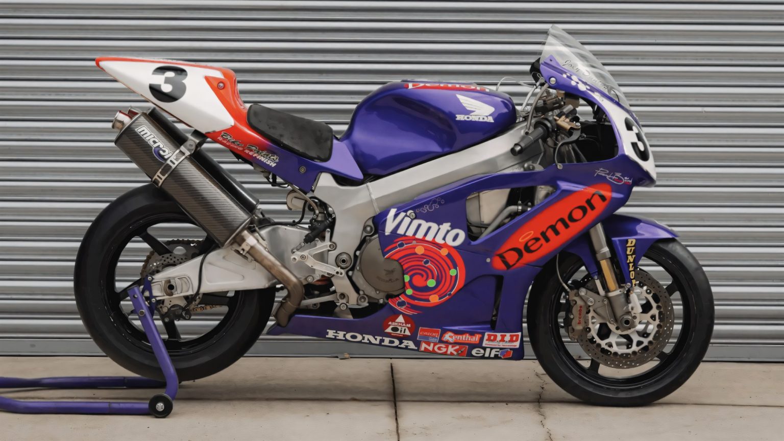 Vimto-Honda SP-1: Joey Dunlops letzte TT-Siegermaschine wird versteigert Lila-weiße Honda VTR1000 SP-1 mit Startnummer 3 und Vimto-Demon-Dekor, fotografiert seitlich vor einem Rolltor bei Iconic Auctioneers