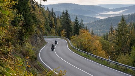 Zwei Motorradfahrer auf Tourenmotorrädern mit Seitenkoffern fahren auf einer kurvigen Bundesstraße mit Leitplanke durch herbstlichen Mischwald im Mittelgebirge, im Hintergrund bewaldete Hügel und Nebel in den Tälern