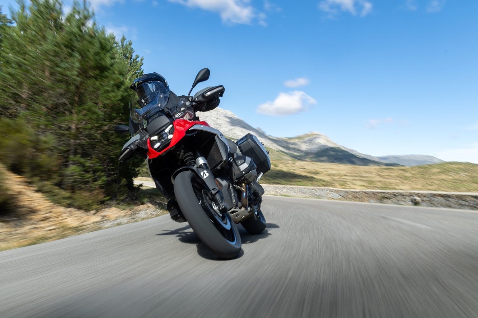 Bridgestone Battlax Adventure A42 – rot-schwarze Reiseenduro mit Koffern in Schräglage auf einer sonnigen Landstraße vor Bergkulisse