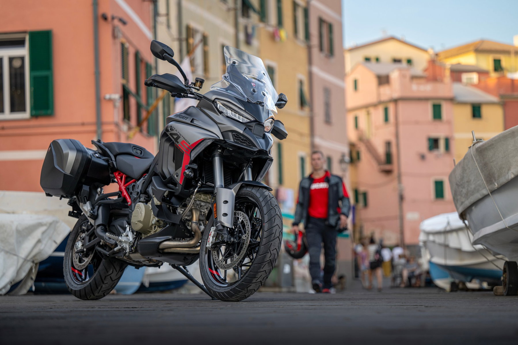 Ducati Multistrada V4 S Grand Tour: The touring Ducati that lowers itself 1 Ducati Multistrada V4 S Grand Tour 2027 – mit schwarzen Seitenkoffern in einer italienischen Hafengasse, im Hintergrund ein Fahrer mit Helm in der Hand