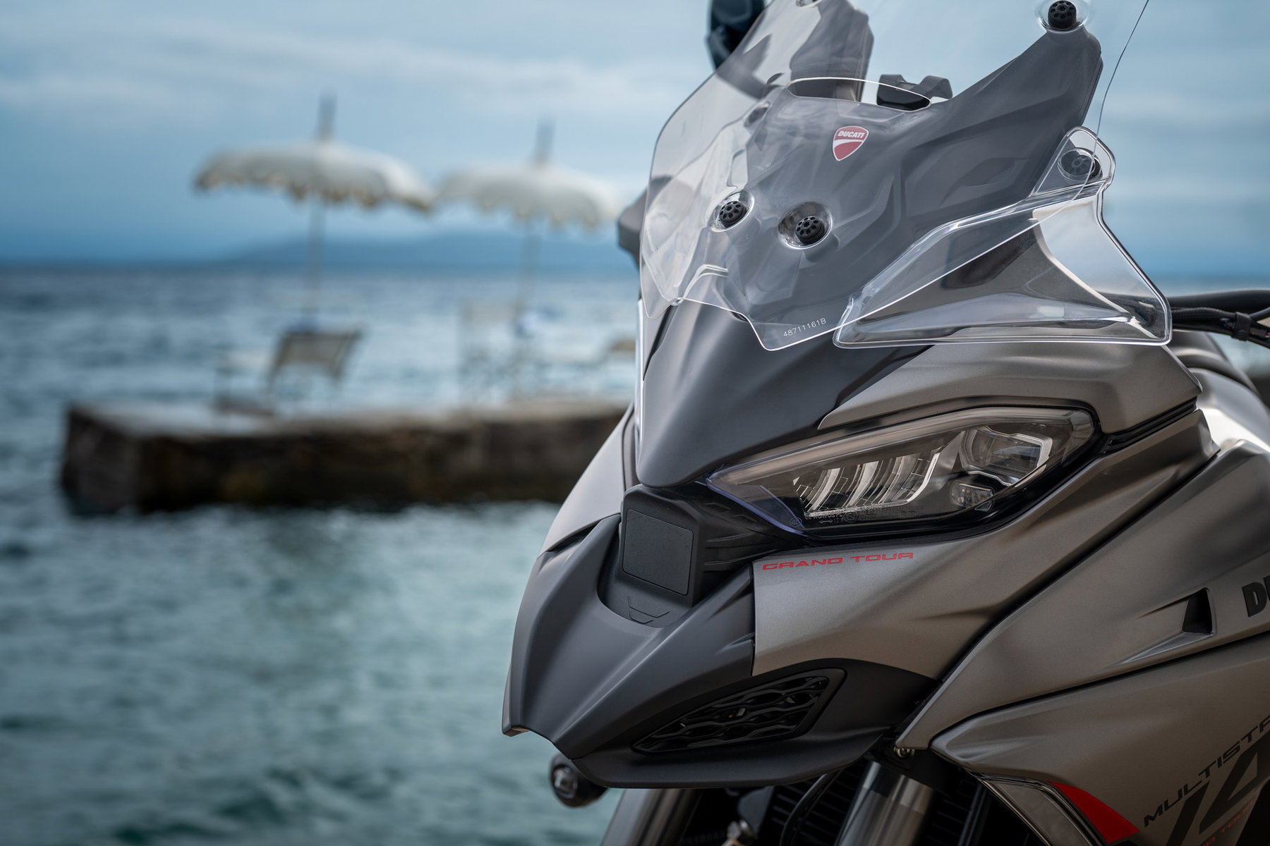 Ducati Multistrada V4 S Grand Tour: The touring Ducati that lowers itself 4 Ducati Multistrada V4 S Grand Tour 2027 – Detailansicht der Front mit LED-Scheinwerfer, Windschild und Grand-Tour-Schriftzug an der Verkleidung vor dem Meer
