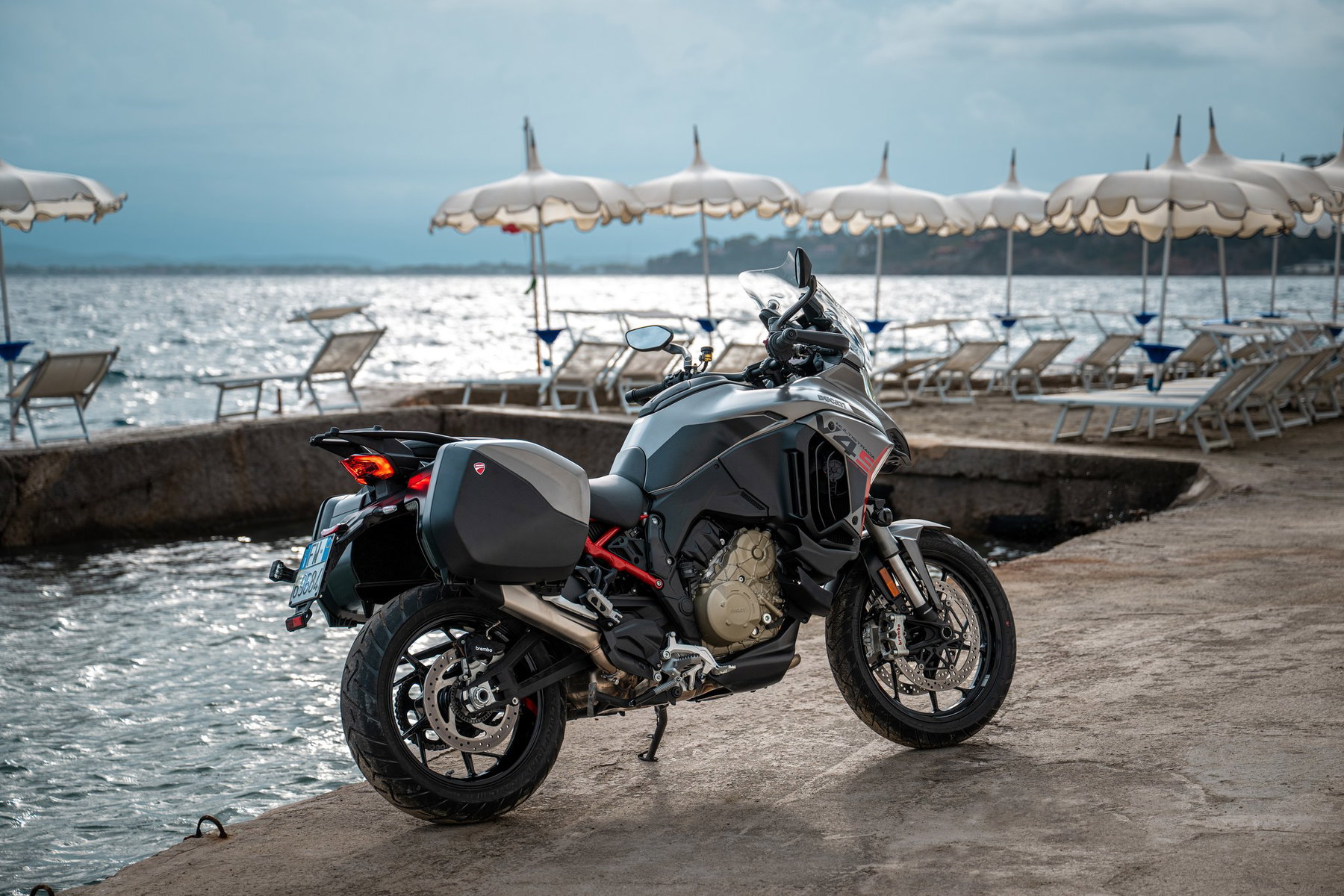 Ducati Multistrada V4 S Grand Tour: The touring Ducati that lowers itself 2 Ducati Multistrada V4 S Grand Tour 2027 – Heckansicht schräg von links mit Seitenkoffern, roten Akzenten und V4-Motor auf einer Mole am Strand