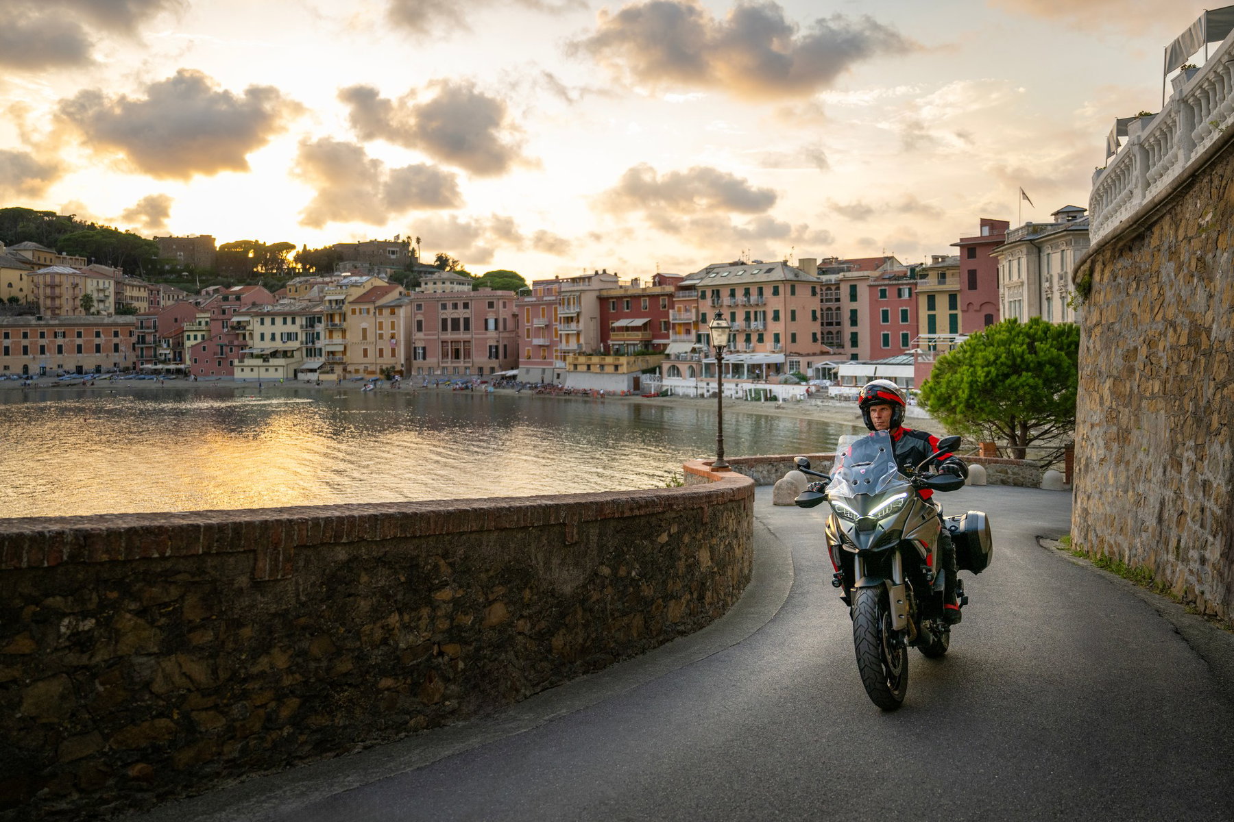 Ducati Multistrada V4 S Grand Tour: The touring Ducati that lowers itself 5 Ducati Multistrada V4 S Grand Tour 2027 – Fahraufnahme auf einer Uferstraße bei Sonnenuntergang vor einem ligurischen Küstenort