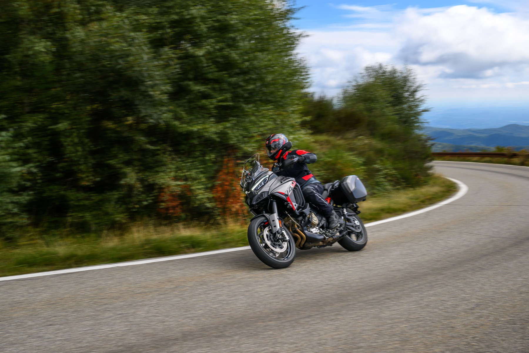 Ducati Multistrada V4 S Grand Tour: The touring Ducati that lowers itself 3 Ducati Multistrada V4 S Grand Tour 2027 – Fahraufnahme in Schräglage in einer Kurve mit Blick aufs Meer, mit Seitenkoffern