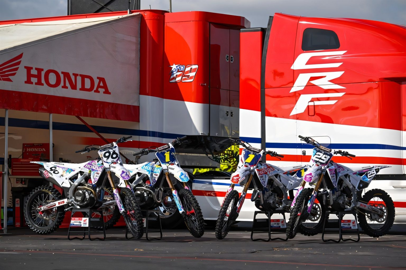 Signed Honda Factory Parts From 30 Dollars: Auction Helps Children With Brain Tumors Mehrere Honda CRF-Werksmotorräder in weißer Sonderlackierung mit bunten Kinder-Motiven vor dem roten Honda-Truck