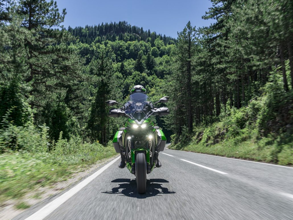Kawasaki Versys 900: Z900 four-cylinder and radar close the Versys gap 2 Kawasaki Versys 900 SE 2027 frontal auf einer Landstraße im Wald mit eingeschalteten LED-Scheinwerfern und Nebelscheinwerfern