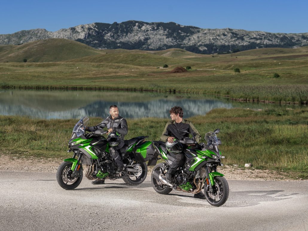Kawasaki Versys 900: Z900 four-cylinder and radar close the Versys gap 5 Zwei Fahrer auf Kawasaki Versys 900 SE 2027 in Plasma Lime Green stehen mit Koffern an einem Bergsee vor felsiger Hügellandschaft