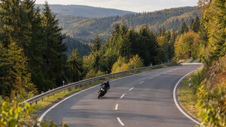 Motorradfahrer in Schräglage auf breiter, kurviger Bundesstraße mit Leitplanke durch herbstlich bewaldetes Mittelgebirge – Symbolbild zur Lützel (B62) im Siegerland