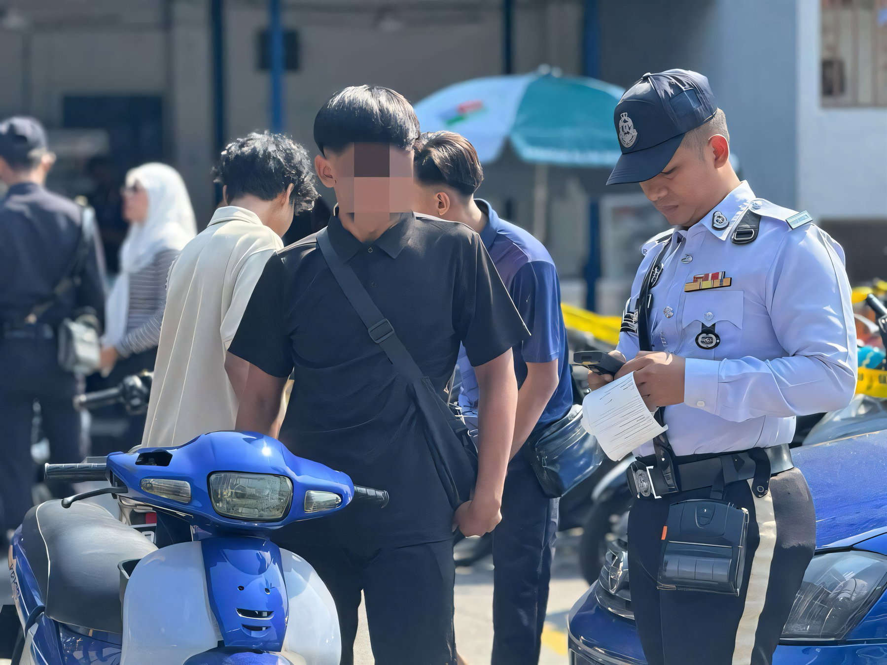 Mat Rempit in Penang: 30 Motorräder kassiert, ab November droht Haft 5 Ein Beamter der malaysischen Verkehrspolizei JSPT stellt bei einer Kontrolle einen Strafzettel aus, daneben junge Motorradfahrer mit verpixeltem Gesicht und ein blauer Roller