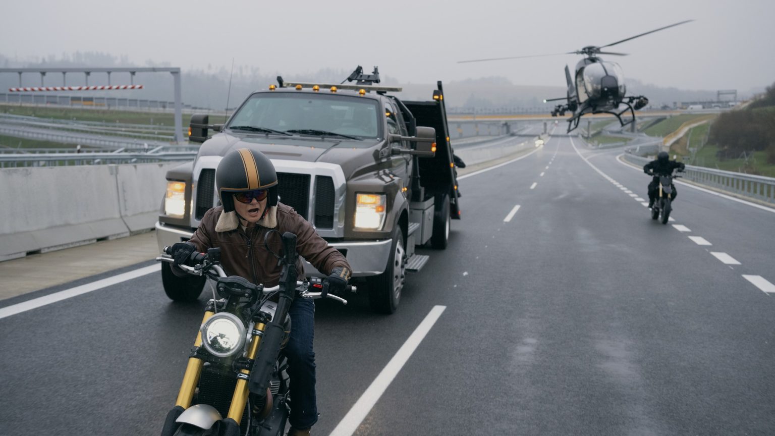 Triumph Scrambler 1200 XE in Matchbox The Movie – Fahrer mit Jethelm und Lederjacke auf nasser Autobahn, verfolgt von Abschleppwagen, Hubschrauber und zweitem Motorrad
