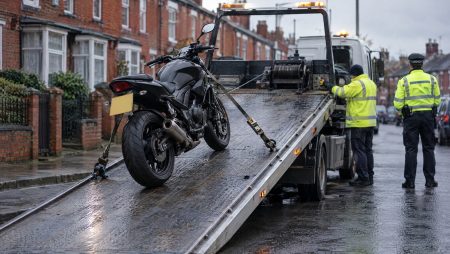 Schwarzes Naked Bike wird in einer englischen Reihenhausstraße auf einen Abschleppwagen gezogen, daneben ein Polizist und ein Abschlepper in Warnjacken bei Regenwetter