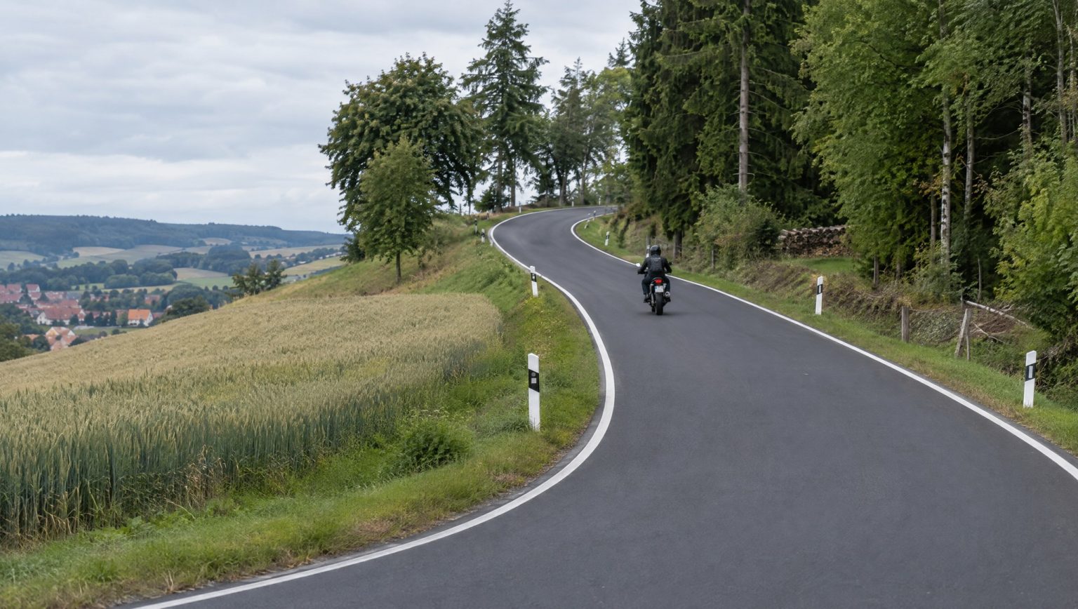 St2190 Katschenreuth – Krumme Fohre: Zweiradsperre nach Sanierung aufgehoben – Überblick & Chronik Motorradfahrer auf einer frisch asphaltierten, kurvigen Landstraße zwischen Getreidefeld und Waldrand in Oberfranken, im Tal ein Dorf, bewölkter Himmel