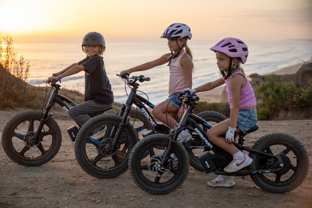 Drei Kinder auf elektrischen Stacyc-Laufrädern 16eDrive und 20eDrive bei Sonnenuntergang über dem Meer