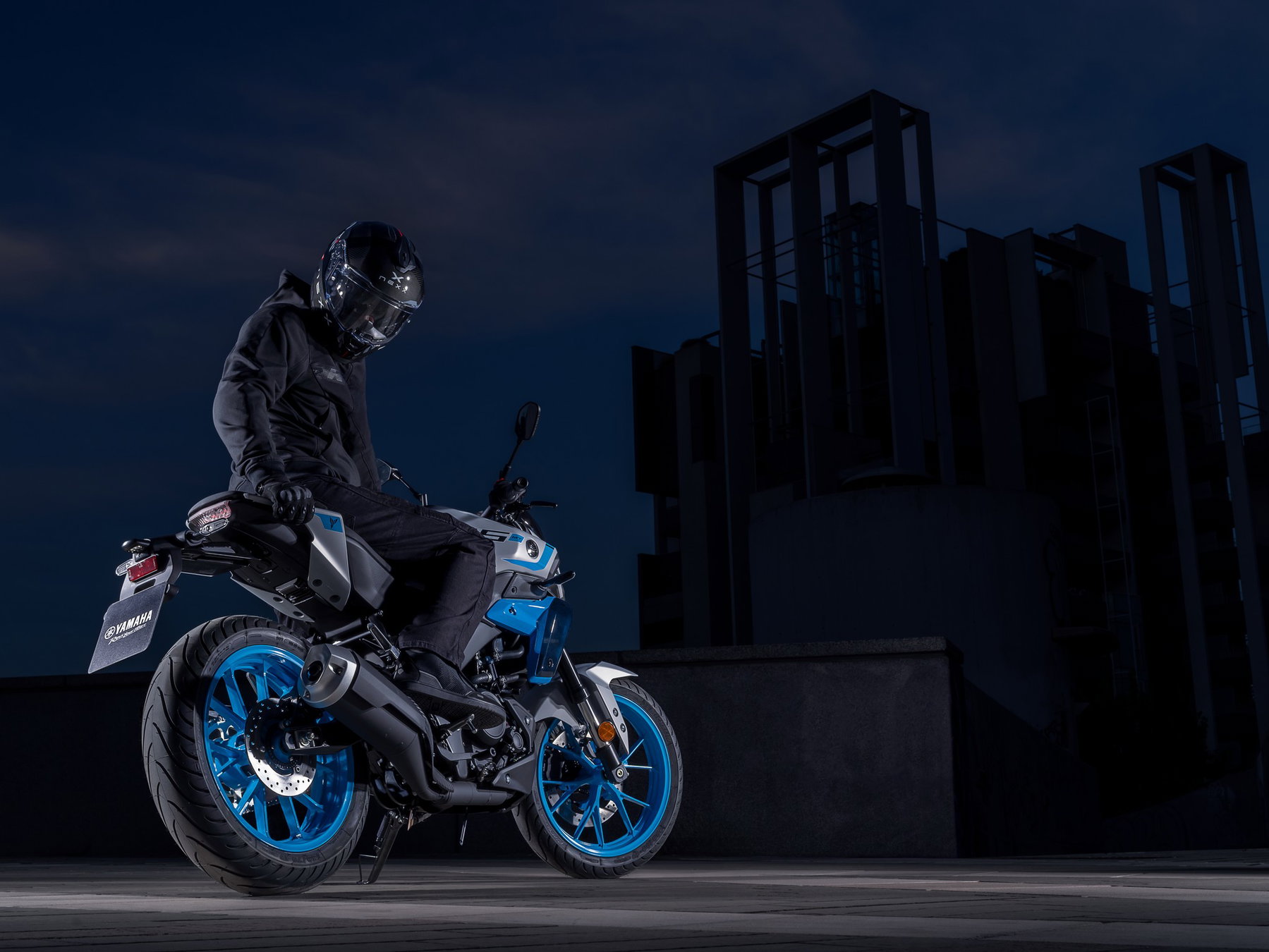 Yamaha MT-125 2027 – Fahrer sitzt auf der MT-125 in Ice Storm (Silbergrau mit hellblauen Felgen) vor nächtlicher Stadtkulisse