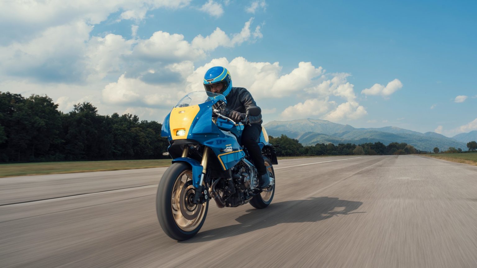 Yamaha XSR900 GP 2027 – Fahraufnahme in Legend Blue mit gelber Startnummerntafel auf einer Flugplatz-Piste vor Wolkenhimmel