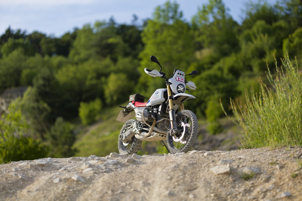 BMW R 12 G/S GS Trophy Competition Bike 2026 – Frontansicht in Lightwhite mit Startnummer 123 auf Schotterkuppe vor grünem Wald