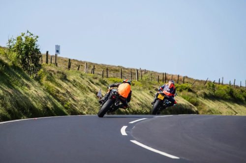Zwei Newcomers-Solofahrer von hinten in Schraege auf dem Mountain Course – Isle of Man TT 2026 Speed Controlled Lap gruene Huegel und Weidezaeune