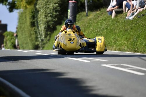 Sidecar Nr 37 Ellum Mannan Joinery SMH Racing von hinten durch ein Dorf – gelbes Gespann Isle of Man TT 2026 Newcomers Speed Controlled Lap Baeume und Zuschauer