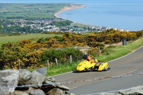 Gelbes Sidecar-Gespann auf der Bergstrecke des Mountain Course – Isle of Man TT 2026 Newcomers mit Blick auf Dorf und Irische See