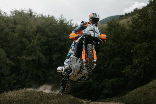KTM 890 Adventure R Rally 2026 beim Sprung ueber Erdhügel – Frontansicht Offroad bewoelkter Himmel