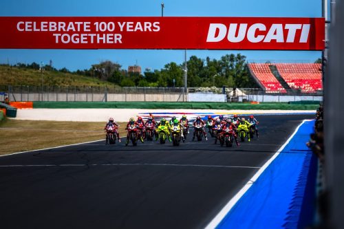 Gruppe von Ducati-Werksfahrern beim Start auf der Strecke unter dem Celebrate-100-Years-Banner in Misano