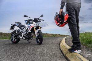 BMW F 450 R 2027 – M Sport in Snaefell Weiß / M Motorsport, Standaufnahme schräg von vorn rechts, daneben Fahrer mit rotem Helm in der Hand