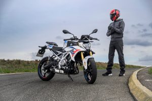 BMW F 450 R 2027 – M Sport in Snaefell Weiß / M Motorsport, Standaufnahme schräg von vorn rechts, daneben Fahrer mit rotem Helm