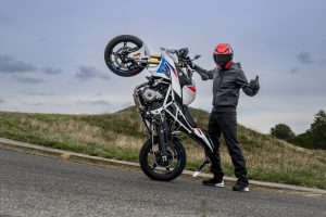 BMW F 450 R 2027 – M Sport in Snaefell Weiß / M Motorsport auf dem Hinterrad aufgestellt, daneben Fahrer mit rotem Helm und Daumen nach oben