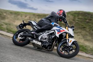 BMW F 450 R 2027 – M Sport in Snaefell Weiß / M Motorsport in tiefer Schräglage, Fahrer mit Knie am Boden auf einer hügeligen Strecke