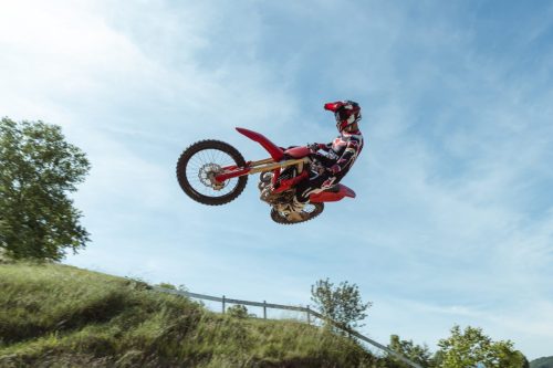 Ducati Desmo250 MX 2027 in Ducati Red – Fahrer springt mit angezogener Maschine über einen Motocross-Hügel vor blauem Himmel, Untersicht auf Showa-Gabel und Vorderrad