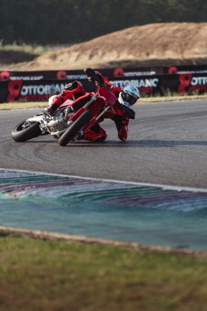 Ducati Desmo450 SM 2027 – Schräglage auf der Rennstrecke vor Offroad-Hügel, Hochformat