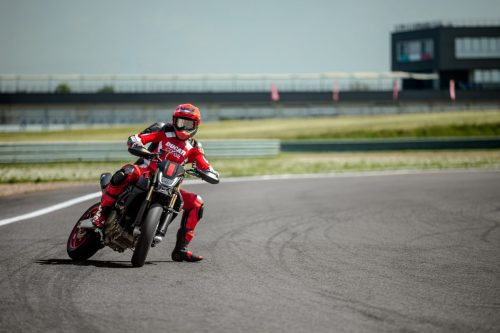 Ducati Hypermotard 698 Mono Nera 2027 – Fahrer in roter Ducati-Corse-Kombi beim Supermoto-Drift in Schräglage auf Rennstrecke mit Boxengebäude im Hintergrund