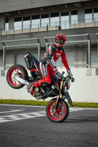 Ducati Hypermotard 698 Mono Nera 2027 – Stoppie-Wheelie frontal auf Rennstrecke, Fahrer in Ducati-Corse-Kombi, rote Gussräder und schwarze Lackierung sichtbar