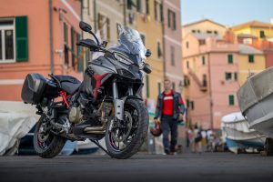Ducati Multistrada V4 S Grand Tour 2027 – mit schwarzen Seitenkoffern in einer italienischen Hafengasse, im Hintergrund ein Fahrer mit Helm in der Hand