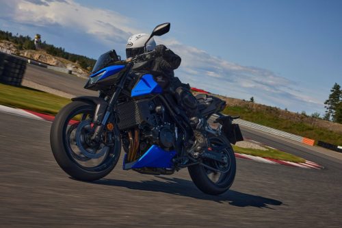 Honda CB750 Hornet 2027 in Glint Wave Blue in Schräglage auf einer Rennstrecke, Fahrer mit weißem Helm und schwarzer Lederkombi, Frontansicht von schräg links
