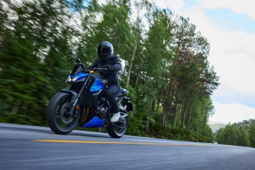 Honda CB750 Hornet 2027 in Glint Wave Blue auf einer Landstraße im Wald, Fahrer in Alltagskleidung, Frontansicht von schräg links in Bewegung