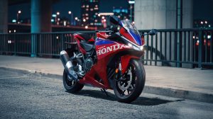 Honda CBR500R FOUR 2027 in Ignition Red Tricolour nachts unter einer Brücke vor der Stadtkulisse, Dreiviertelansicht von vorne